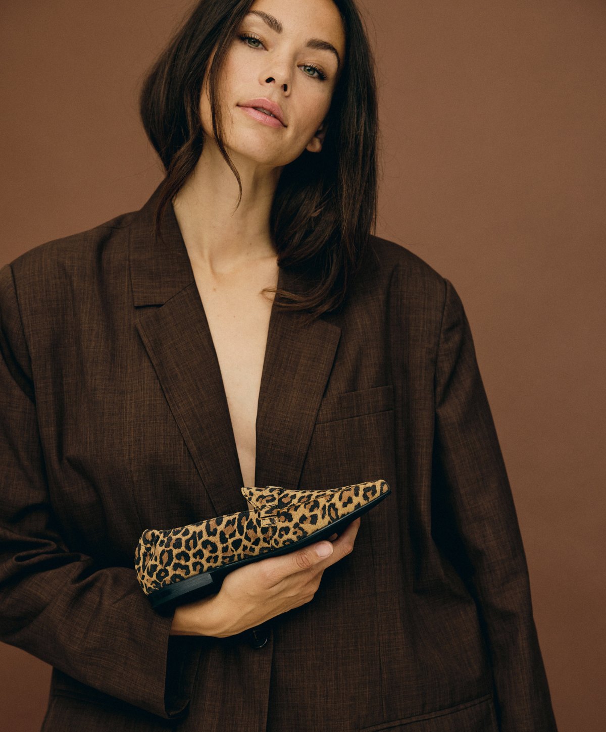 Kvinde iført en stor brun jakke fra Shoedesign Copenhagen, holder en leopardmønstret sko fra samme mærke foran en brun baggrund.