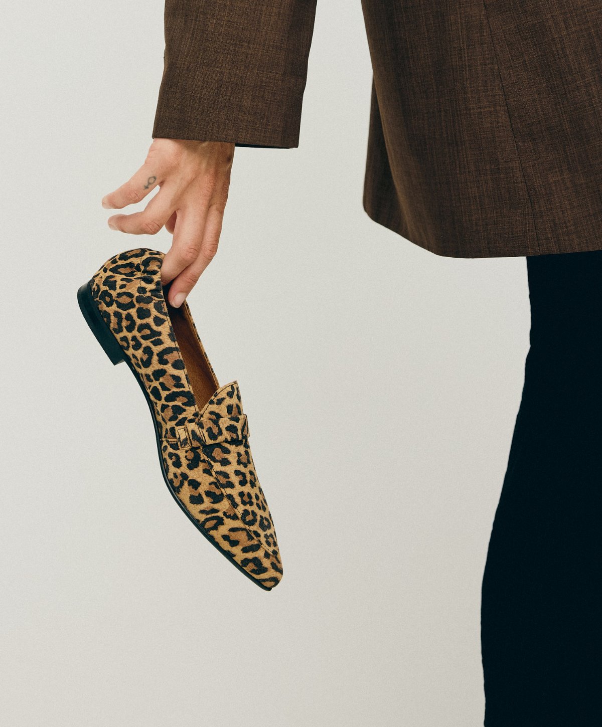 En person iført en brun jakke og sorte bukser holder en leopardmønstret sko fra Shoedesign Copenhagen.