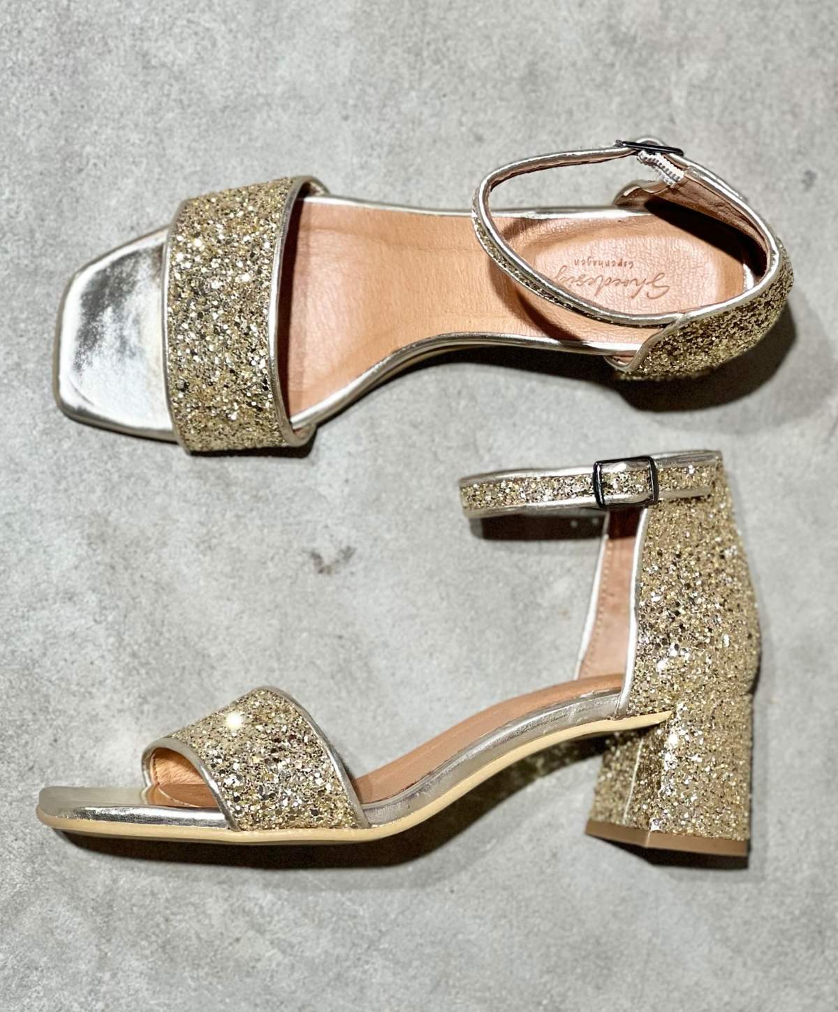 Guldglitrende højhælede sandaler fra Shoedesign Copenhagen med ankelrem og blokhæl, set ovenfra på en grå baggrund.
