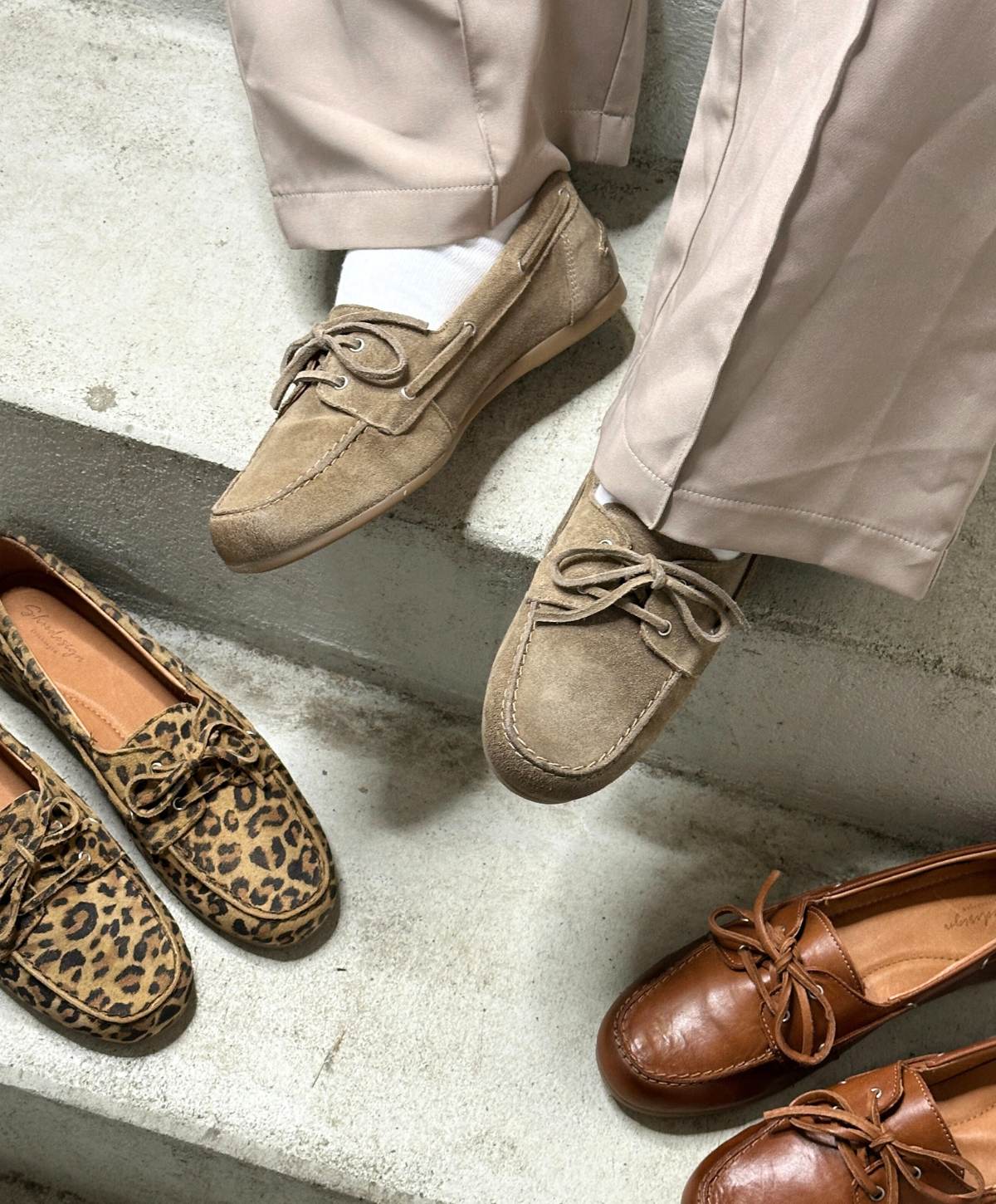Person iført beige bukser og lysebrune mokkasiner fra Shoedesign Copenhagen, sidder på en trappe. Ved siden af står et par leopardmønstrede og et par brune mokkasiner fra samme mærke.