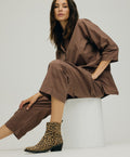 Kvinde iført brun jumpsuit og leopardmønstrede ankelstøvler fra Shoedesign Copenhagen, sidder på en hvid cylinderformet skammel.