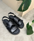 Sort læder sandal fra Shoedesign Copenhagen med krydsede remme og justerbar spænde, placeret på en betonoverflade ved siden af grønne blade.