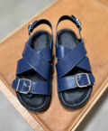 Blå lædersandaler med krydsremme og sølvspænder fra Shoedesign Copenhagen, placeret på en brun overflade.