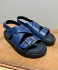 Blå sandaler fra Shoedesign Copenhagen med krydsede remme og sølvspænder, placeret på en brun overflade.