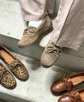 Person iført beige bukser og lysebrune mokkasiner fra Shoedesign Copenhagen, sidder på en trappe. Ved siden af står et par leopardmønstrede og et par brune mokkasiner fra samme mærke.