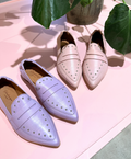 To par sko fra Shoedesign Copenhagen i pastelfarver, en i lilla og en i lyserød, med nitter og dekorative syninger, placeret på en lyserød overflade ved siden af en grøn plante.